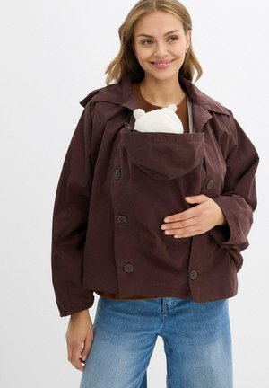 Giovane donna che indossa un cappotto marrone con un marsupio incorporato, tiene un orsacchiotto bianco, sorridendo, su uno sfondo semplice.