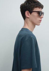 Maglietta a maniche corte color verde petrolio scuro, realizzata in tessuto morbido, con vestibilità comoda e dettagli di cuciture visibili. Indossata con occhiali da sole a motivo.