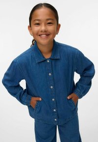 Denim jaka vidējā zilā krāsā, ar pogām frontē, apkakli un elastīgiem aprocēm, kombinēta ar atbilstošām biksēm, lai izveidotu pilnīgu izskatu.