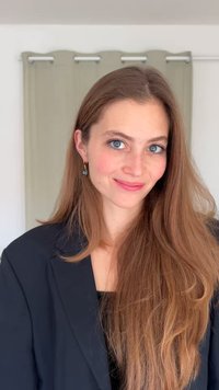 Μαύρο σακάκι, ελαφρύ ύφασμα, μακριά καστανά μαλλιά, μπλε μάτια, φοράει χρυσά σκουλαρίκια με μπλε λεπτομέρεια. Απαλό φόντο με φωτεινές κουρτίνες.