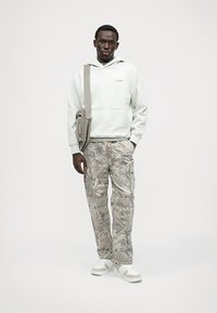 Jasnoszara bluza z kapturem z przednią kieszonką, w połączeniu z wzorzystymi spodniami cargo w odcieniach szarości. Białe sneakersy dopełniają stylizację.