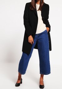 Svart lång kappa över en krämfärgad stickad tröja, kombinerad med blå vida denimjeans. Modellen har också på sig svarta högklackade skor.