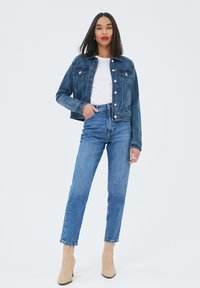 Veste en denim foncé avec des boutons en métal, deux poches poitrine et une coupe courte. Associée à un jean bleu taille haute et des bottines beige.