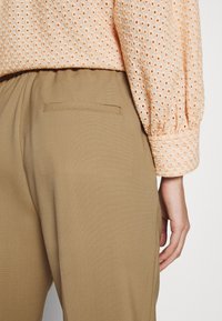 Perzikkleurige blouse met perforaties en pofmouwen, gecombineerd met beige broeken met een achterzak en een gladde textuur.