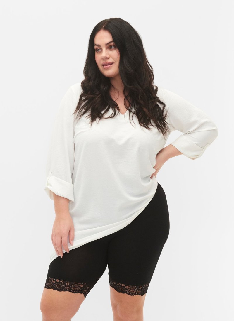 Pitkähiuksinen nainen, joka pukeutuu valkoiseen pitkähihaiseen paitaan ja mustiin pitsireunaisiin pyöräilyshortseihin, seisoo käsi lanteilla.