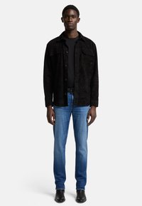 Giacca a camicia in suede nero, maniche lunghe, con due tasche sul petto, abbinata a jeans scuri e stivali in pelle nera. T-shirt nera semplice sotto.