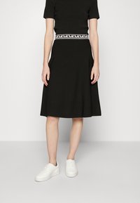 Abito nero con maniche corte, silhouette svasata e una fascia in vita a motivi bianchi. Abbinato a sneaker bianche. Sfondo semplice.