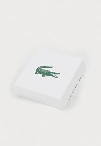 Baltas stačiakampis langelis su tekstūruota paviršiumi, ant viršaus esantis žalias krokodilo logotipas ir šone užrašas „LACOSTE“ žalia spalva.