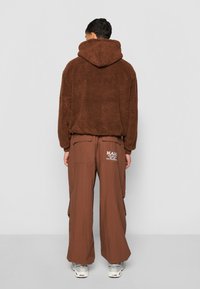 Brun fleece hoodie med rund hætte, parret med løstsiddende brune cargo bukser med lommer og logo detaljer på ryggen.