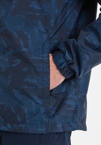 Navy blue windbreaker with a textured abstract pattern, elastic cuffs, and side pockets. Smooth, lightweight material.