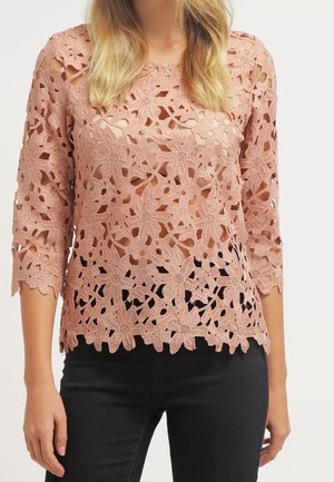 Mujer con un top de encaje de crochet rosa polvoriento con patrones florales y mangas tres cuartos, combinado con pantalones negros.