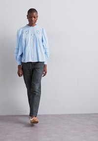 Lys blå, langermet bluse med pufferm og broderte detaljer, kombinert med mørke jeans og lysebrune slip-on sko.