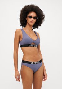 Fialový žebrovaný bikinový set s vykrojeným designem a širokým černým páskem s logem "CALVIN KLEIN". Texturovaný materiál, elegantní tvar.