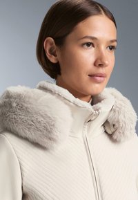 Világos bézs dzseki puha, világos szőrmés gallérral. Az anyag átlós, steppelt mintázattal rendelkezik, és elöl cipzárral záródik.