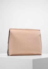 Beige kožená kabelka s obdélníkovým tvarem a minimálním prošíváním, umístěná na bílém geometrickém podstavci na jednobarevném pozadí.