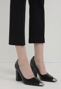 Scarpe nere con tacco alto e punte argentate indossate con pantaloni neri a cropped su uno sfondo neutro.