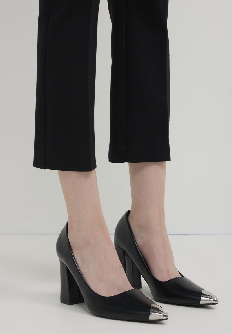 Scarpe nere con tacco alto e punte argentate indossate con pantaloni neri a cropped su uno sfondo neutro.