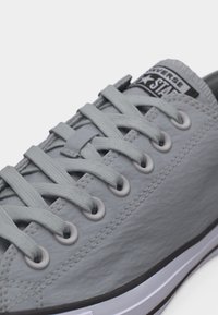 Κοντινό πλάνο ενός γκρι αθλητικού παπουτσιού Converse, με έμφαση στα κορδόνια, τις τρύπες, τις ραφές και το λογότυπο στη γλώσσα του παπουτσιού.