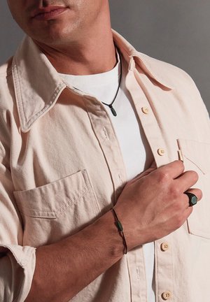 Light pink corduroy shirt over a white t-shirt. Black necklace, green bracelet, and hexagonal black ring on the hand. Close-up of details.
