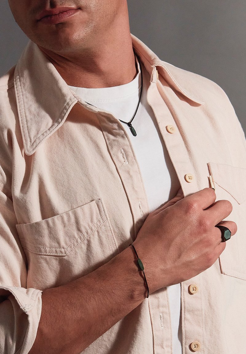 Lyserød corduroy skjorte over en hvid t-shirt. Sort halskæde, grøn armbånd og sekskantet sort ring på hånden. Nærbillede af detaljer.