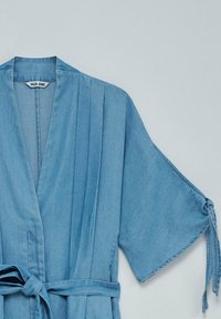 Hellblauer Denim-Mantel mit Bindegürtel und weiten Ärmeln, mit vertikalen Falten entlang der Vorderseite. Das Etikett trägt die Aufschrift "Salsa Jeans."