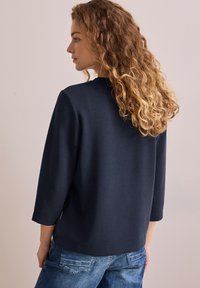 Marineblaues Oberteil mit drei Viertelärmeln aus weichem Stoff und runder Halslinie. Kombiniert mit hellblauen, figurbetonten Jeans.
