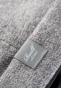 Tela de tejido textil gris con una trama texturizada, que presenta una etiqueta rectangular con "Reell" en cursiva blanca sobre un fondo gris claro.