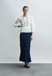 Blouse blanche plissée à manches trois-quarts ; jupe marine superposée évasée ; chaussures plates noires ; collier simple en argent.