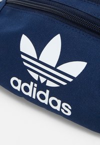 Mörkblå, strukturerad tygväska med en vit Adidas-logotyp som har tre bladliknande accenter och tre horisontella ränder. Dragkedjans stängning är synlig.