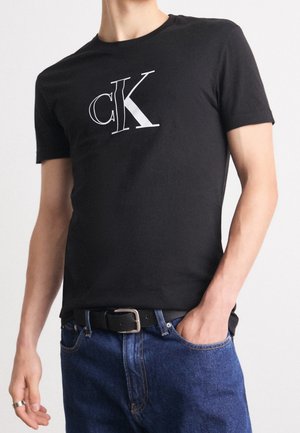 Personne portant un t-shirt noir ajusté à manches courtes avec un grand logo blanc « cK », un jean bleu, une ceinture noire, une bague argentée, la main dans la poche.