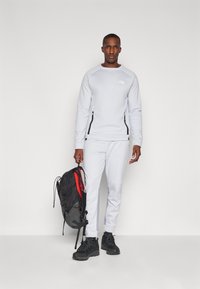 Sweatshirt cinza claro com material elástico, zíperes laterais e logótipo. Calças cinzas a condizer, sapatos de caminhada pretos e uma mochila preta com destaques vermelhos.