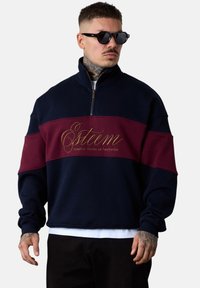 Sweat-shirt coloré en blocs marine et bordeaux avec un col haut, une fermeture éclair et un logo brodé en or. Tissu doux, coupe décontractée, poignets côtelés.