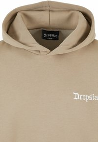 Beiger Kapuzenpullover mit Kordelzug, weichem Material und einem Frontlogo in weißer Schreibschrift. Innen am Kragen mit "Dropsize – groß" etikettiert.