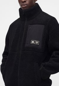 Homme portant une veste polaire noire zippée avec un tissu texturé et une poche poitrine zippée avec un écusson du logo Kappa.