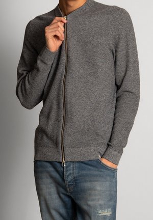 Mężczyzna w szarym swetrze zapinanym na suwak o teksturowanej powierzchni, jedna ręka podciągająca zamek, druga w kieszeni, noszący niebieskie dżinsy z przetarciami i wyblakłymi miejscami.