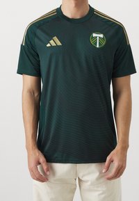 Henkilö, jolla on yllään tummanvihreä jalkapallopaita, jossa on kultainen Adidas-logo ja Portland Timbersin vaakuna, yhdistettynä vaaleanbeigeihin housuihin.