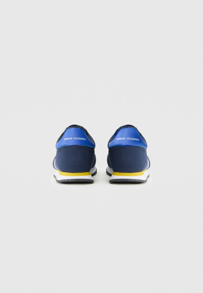 Zapatillas deportivas azul marino con una superficie texturizada, media suela blanca y un acento amarillo. Presentan la marca "ARMANI EXCHANGE" en el talón.