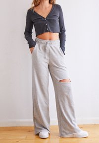 Kvinna iförd en mörkgrå kort knappad topp, ljusgrå vida byxor med ett knäutskärning och vita sneakers, stående på ett trägolv.