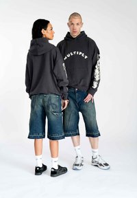Sort hættetrøje med "MULTIPLY" tekst og flammegrafik; parret med løse, slidte denimshorts. Modellens fodtøj: sorte sneakers, hvide sokker.