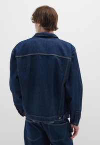 Giubbotto in denim blu scuro con vestibilità comoda. Presenta colletto classico, polsini con bottoni, cuciture a vista e dettaglio della cucitura a yoke sul retro.