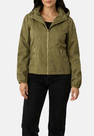 Donna che indossa una giacca con cappuccio verde oliva con cerniere laterali e polsini elastici, abbinata a pantaloni neri.