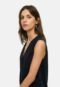 Mujer con cabello largo y castaño, que lleva una camiseta negra sin mangas y pendientes colgantes, mirando ligeramente de lado sobre un fondo claro.