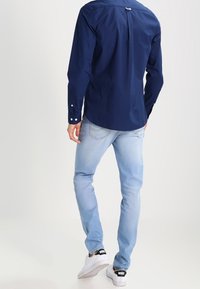 Camisa de botones azul marino con un pliegue en la parte de atrás, combinada con jeans azul claro y zapatillas blancas con acentos negros. Tejidos suaves y corte ajustado.