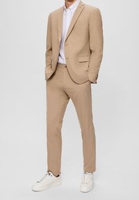 Beige jakkesæt lavet af glat stof, med en skræddersyet pasform, to-knaps jakke og flade bukser, parret med hvide sneakers.