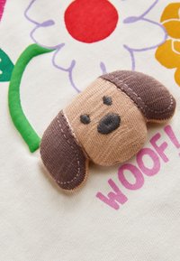 Pieni kangaskoiran pää, jossa on ruskeat korvat, ommeltu valkoiseen kankaaseen, jossa on värikkäitä kukkia ja sana "WOOF!" vaaleanpunaisilla kirjaimilla.
