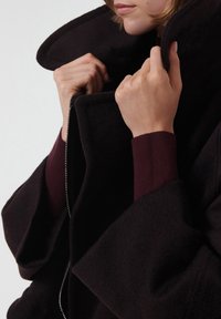 Abrigo de lana negro con un cuello alto, con una cremallera plateada y puños de punto en color burdeos. La tela parece suave y texturada.