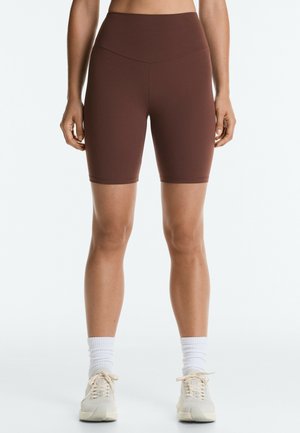 Femme portant un cuissard marron taille haute, des chaussettes blanches et des chaussures de sport beiges, debout devant un fond clair uni.