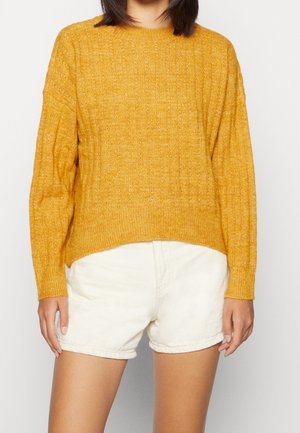 Person iført en sennepsgul strikket sweater og hvide shorts med høj talje, stående mod en ensfarvet lys baggrund.