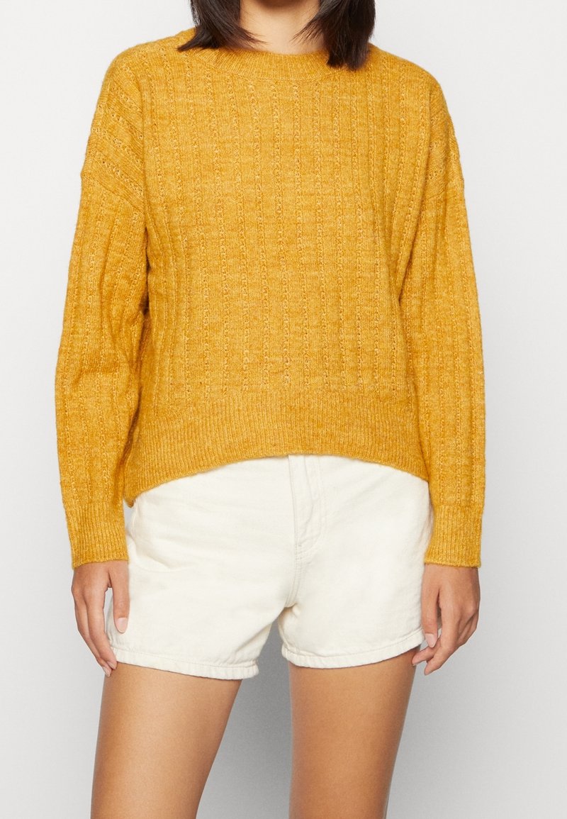 Personne portant un pull en tricot jaune moutarde et un short blanc taille haute, debout devant un fond clair uni.
