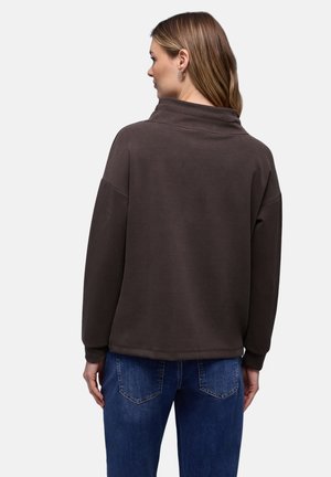 Ruskea collegepaita, jossa on korkea kaulus, rento istuvuus ja lasketut olkapäät. Yhdistetty sinisiin farkkuihin, joissa on takataskut ja haalistuneet yksityiskohdat.